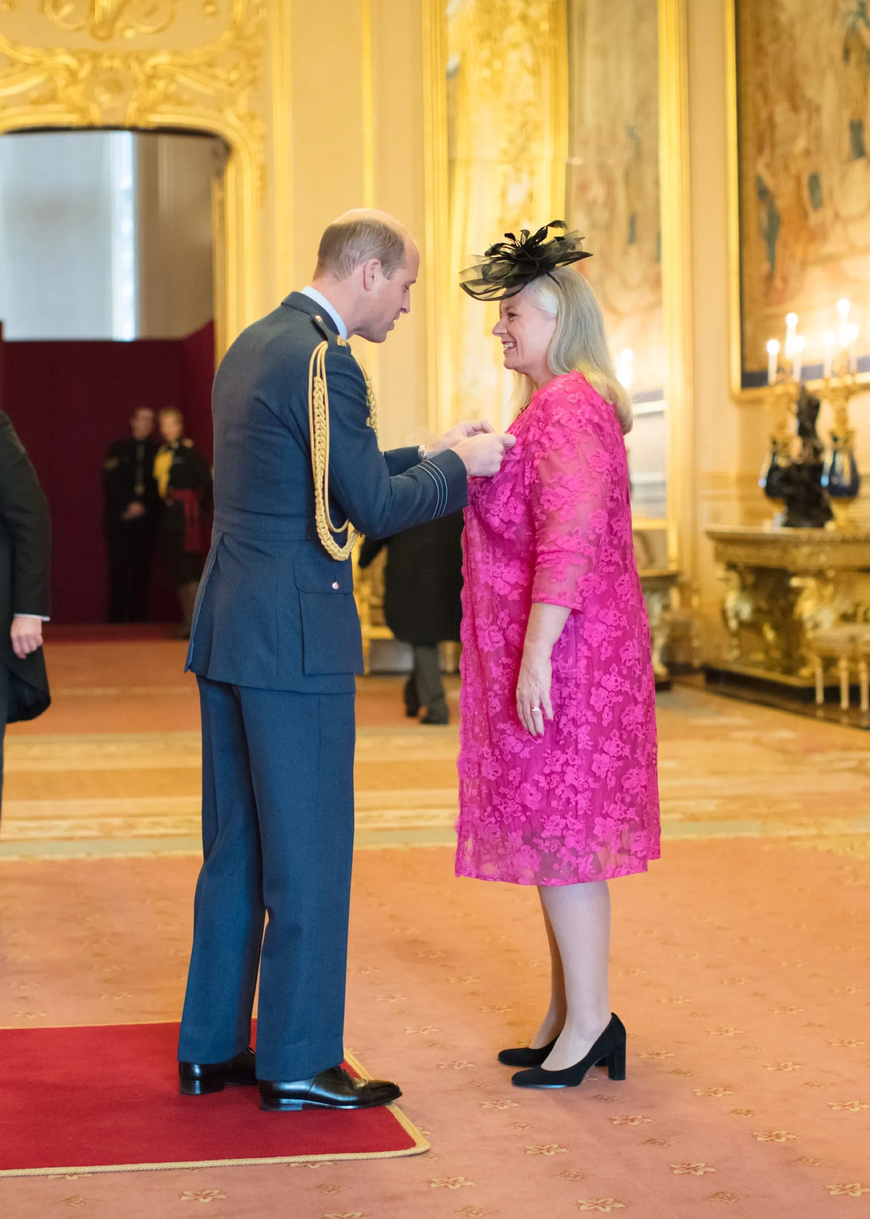 Dr Alison Edgar MBE receiving her MBE from Prince William at Windsor Castle, in a pink lace dress and black fascinator.